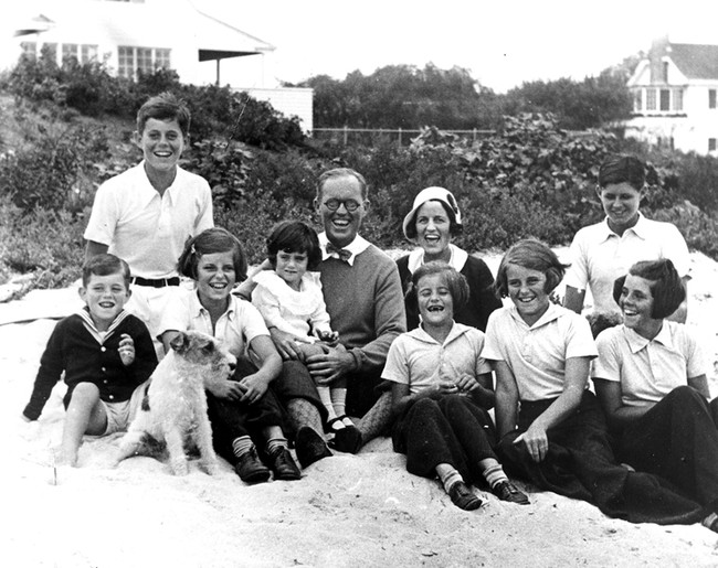 PC 8 The Kennedy Family at Hyannis Port, 1931. L-R: Robert Kennedy, John F. Kennedy, Eunice Kennedy, Jean Kennedy (on lap of) Joseph P. Kennedy Sr., Rose Fitzgerald Kennedy (behind) Patricia Kennedy, Kathleen Kennedy, Joseph P. Kennedy Jr. (behind) Rosemary Kennedy. Dog in foreground is "Buddy". Photograph by Richard Sears in the John F. Kennedy Presidential Library and Museum, Boston.